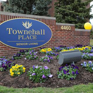 A welcoming sign for Towne Hall Place Senior Living, surrounded by colorful flowers and a brick wall.