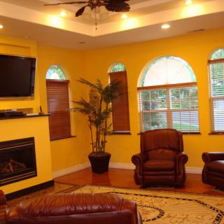A cozy living room with yellow walls features a television mounted above a fireplace, two armchairs, wooden blinds on windows, and a decorative plant.