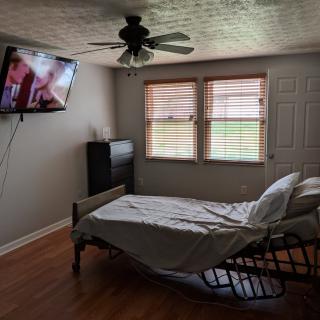 A bedroom with a hospital bed, wall-mounted TV, and windows with blinds.