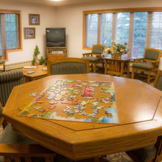 A cozy game room featuring a wooden table with a puzzle, surrounded by comfortable seating and large windows.