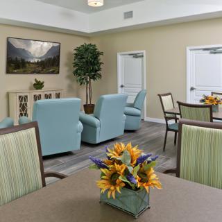 A cozy living room area with light blue armchairs, green striped chairs, tables decorated with flower arrangements, and a landscape painting on the wall.