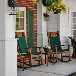 A cozy porch with wooden rocking chairs and green cushions, surrounded by hanging plants and a brick wall.