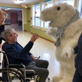 A joyful interaction between an elderly man in a wheelchair and a person dressed in a fluffy animal costume, bringing smiles to the healthcare setting.