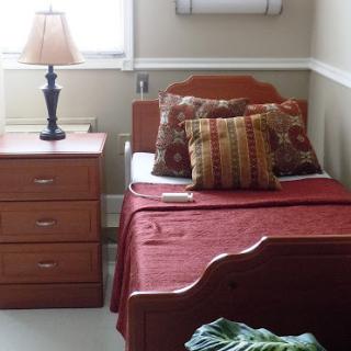 A cozy bedroom featuring a single bed with decorative pillows, a nightstand with a lamp, and a neatly arranged atmosphere.