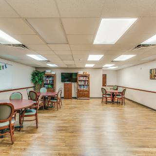 A spacious room with tables, chairs, and a television, featuring wooden flooring and bright lighting.