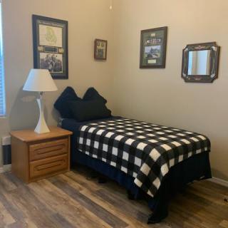 A cozy bedroom with a single bed featuring plaid bedding, accompanied by a nightstand and a lamp, with decorative wall art and a hardwood floor.