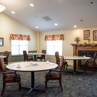 A cozy dining area with tables and chairs, featuring a fireplace and windows with decorative curtains.