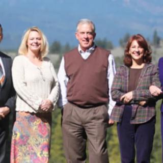 A group of five people standing outdoors and smiling, with a natural background.