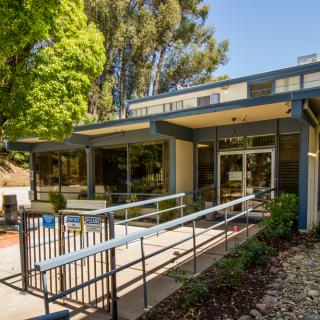 A modern building entrance with a ramp, surrounded by trees and landscaping on a sunny day.