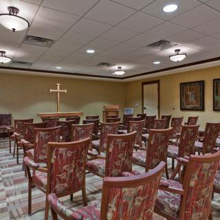 An empty chapel room with arranged chairs, a cross at the front, and warm lighting.