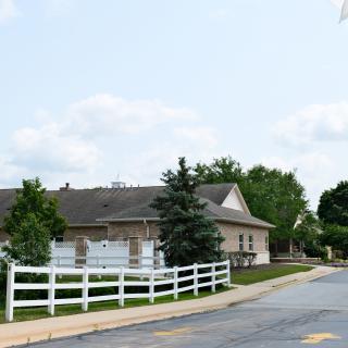 A suburban house with a white fence and trees lining the driveway under a partly cloudy sky.