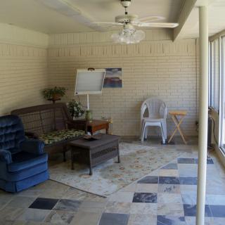 A cozy sunroom featuring a blue armchair, wooden sofa, and coffee table on a patterned rug with tile flooring.