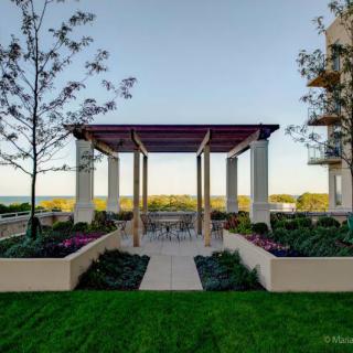 A beautifully landscaped outdoor area featuring a pergola with seating, surrounded by lush greenery and trees.