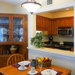 A cozy dining area with a view into the kitchen, featuring a wooden table set for two and adorned with a fruit basket.