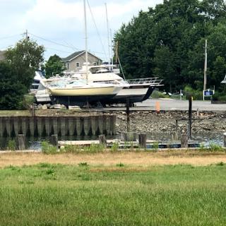 A boat on dry land at a marina, surrounded by grass and trees under a cloudy sky.