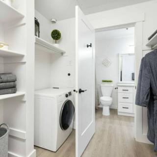 A modern laundry area connected to a bathroom, featuring white walls and shelves with neatly arranged towels.