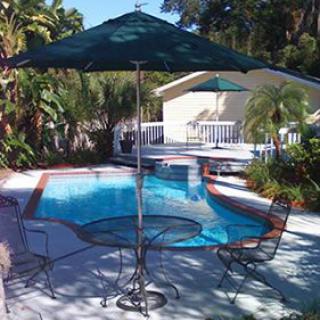 A tranquil outdoor patio with a swimming pool, shaded by an umbrella and surrounded by tropical plants.