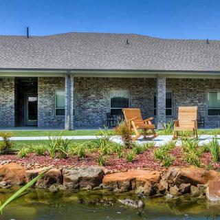 A backyard view featuring a house with patio chairs, a well-maintained lawn, and a picturesque pond with surrounding rocks.