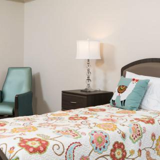 A cozy bedroom featuring a floral patterned bedspread, a bedside table with a lamp, and an upholstered chair.