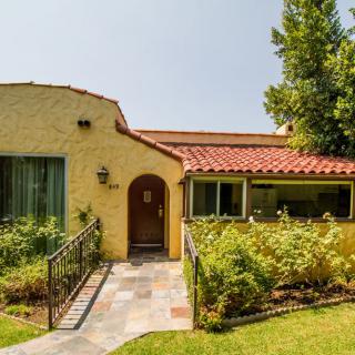 A charming yellow house with a red-tiled roof, surrounded by a lush garden and a neatly paved pathway.