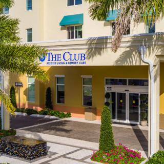 Entrance to a building with 'The Club' sign and surrounding palm trees and landscaping.