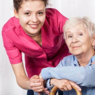 A caregiver in a pink uniform smiles while supporting an elderly woman with a walking aid.
