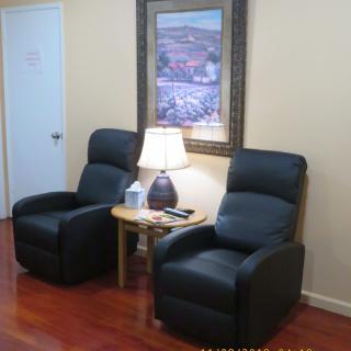 A cozy waiting area with two black recliner chairs, a wooden side table with a lamp, and a painting on the wall.