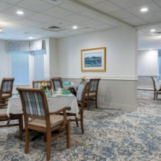 A well-lit dining room with neatly arranged tables and chairs, featuring a patterned carpet and a painting on the wall.