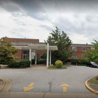 The image shows a building entrance with a driveway, surrounded by trees under a cloudy sky.