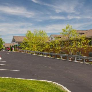 A spacious parking lot in front of a large building surrounded by trees under a blue sky with clouds.
