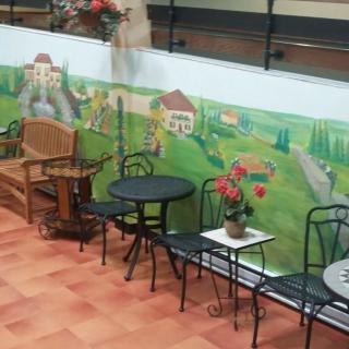 A cozy outdoor seating area featuring a mural of a countryside landscape, complete with tables, chairs, and decorative plants.
