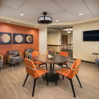 A modern lobby with comfortable seating, featuring a round table surrounded by orange chairs, wall art, and warm lighting.