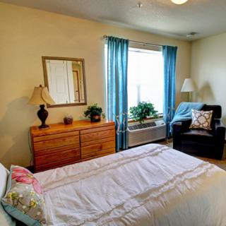 A cozy bedroom with a neatly made bed, a dresser with a mirror, and a comfortable chair by the window.