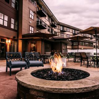 A cozy outdoor patio area with a fire pit surrounded by chairs and an apartment building in the background.
