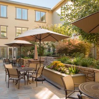 A sunny outdoor patio with tables, chairs, and umbrellas, surrounded by garden landscaping.