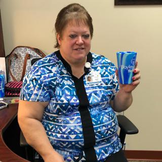A woman in a blue patterned shirt sitting at her desk, happily holding a cup.