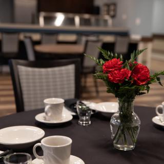 A dining table set with white dishes and a vase of red roses.