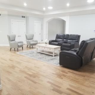 A spacious living room with a minimalist design featuring a black sofa, two grey chairs, a white coffee table, and hardwood flooring.