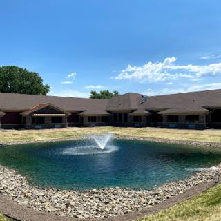 A serene view of a building with a picturesque pond featuring a fountain under a clear blue sky.