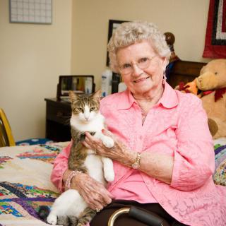 An elderly woman smiling while holding her cat on a bed in a cozy bedroom.
