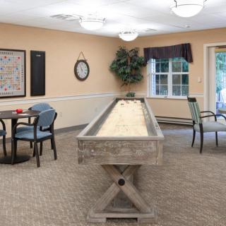 A cozy game room featuring a shuffleboard table, chairs, and a small round table set against carpeted flooring.