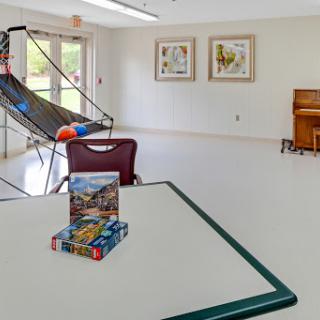 A spacious recreation room featuring a table with puzzles, artworks on the wall, and a piano in the corner.