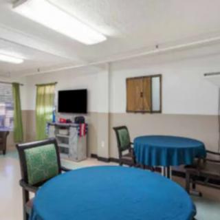 A room with tables and chairs covered in blue cloth, featuring a television and shelves, with curtains on the windows.