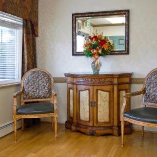 A cozy waiting room with two armchairs and a wooden cabinet topped with a vase of flowers, beneath a wall-mounted mirror.