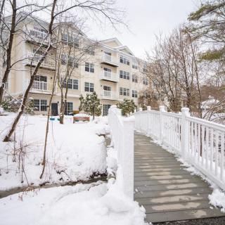 A snowy pathway leading to a multi-story apartment building surrounded by trees.