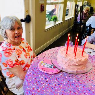 An elderly woman sitting at a table with a pink frosted cake and lit candles, celebrating a birthday.