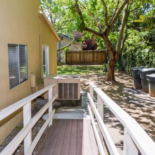 A sunny backyard featuring a wooden ramp leading to a house, with a large tree providing shade.