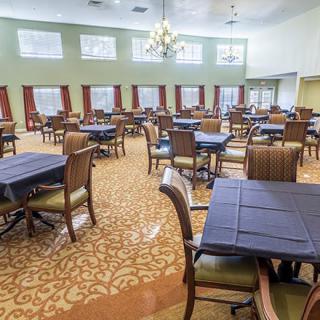 An empty dining hall with several tables and chairs neatly arranged, featuring a chandelier and red curtains.