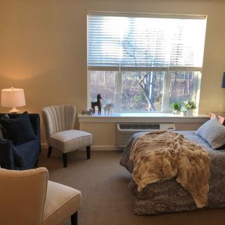 A cozy bedroom interior with a large window, bed, seating area, and soft lighting.