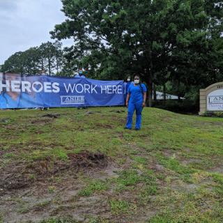 A worker in a blue outfit stands near a large banner that reads 'Heroes Work Here,' set against a grassy background.